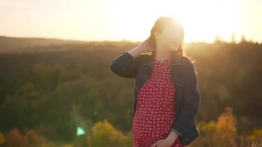Müstakbel anne, genç hamile bir kız güneşin altındaki tepede durup gülümsüyor.. 
