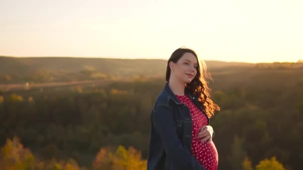 Молода вагітна біла дівчина, що стоїть на пагорбі, торкається її живота на тлі осіннього лісу . — стокове відео