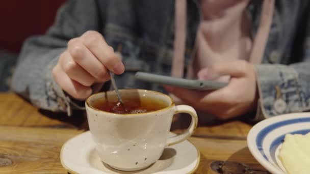 Una chica en una chaqueta de mezclilla revolviendo azúcar en una taza de té usando un teléfono inteligente. — Vídeos de Stock