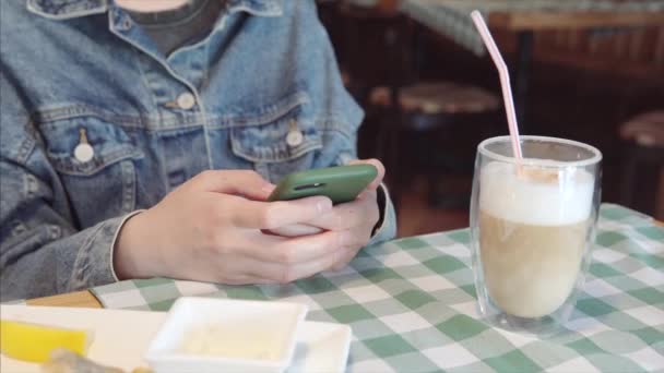 Primer plano de una mujer en chaqueta de mezclilla utiliza teléfono inteligente sentado en un restaurante. — Vídeo de stock
