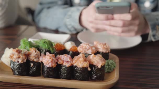 Sushirullar av tonfisk, lax och ål i förgrunden, flickornas händer använder en smartphone i bakgrunden. — Stockvideo