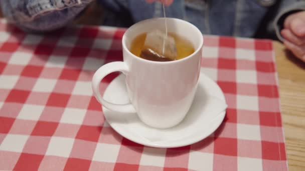 Eine Frau brüht einen Teebeutel in einer weißen Tasse auf einer karierten Tischdecke. — Stockvideo