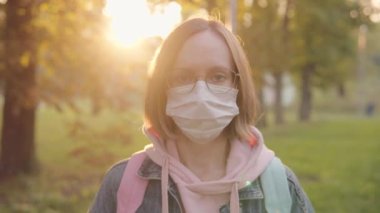 Beyaz koruyucu tıbbi maskeli, gözlüklü, sonbahar parkının arka planında kısa saçlı çekici beyaz kadın.. 