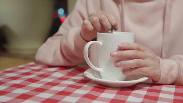 Flickan blandar socker eller stevia med en sked i en keramisk mugg sittande på kvällen i ett café vid bordet. — Stockvideo