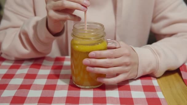 Une fille dans un sweat à capuche rose agite la vitamine thé d'argousier pour les rhumes, boit une boisson chaude réchauffante à travers un gros plan de paille. — Video