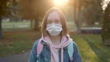 Tıbbi maskeli ve gözlüklü beyaz bir kız akşamları kameraya bakar..