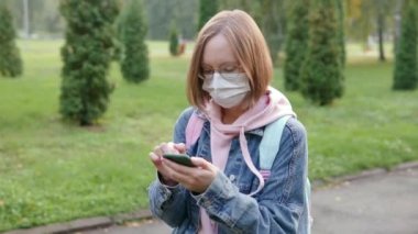 Tıbbi koruyucu maskeli ve gözlüklü güzel bir kadın parkta akıllı telefon kullanıyor, saçları rüzgarda dalgalanıyor. Yavaş çekim.