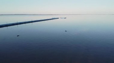 Gün batımında su üzerinde yüzen aşırı gelişmiş sazlıklar ve ördeklerin olduğu gölün yüzeyinin havadan görünüşü. Çok renkli iskelede tatilci insanlar var. Korkmuş bir kuş saklanmaya çalışıyor.