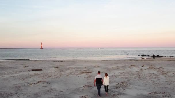 Twee geliefden lopen bij zonsondergang aan de kust met een oude vuurtoren zorgen voor elkaar en knuffelen elkaar. Jong stel op de achtergrond. Een jongen en een meisje. Familie van twee in de buurt van stormachtige kust kijken — Stockvideo