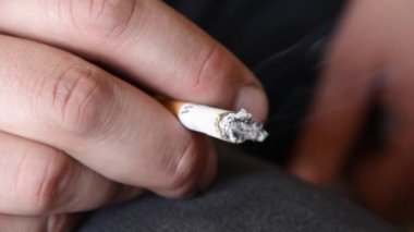 Genç bir adamın elinde sigara içen genç bir adam. Dışarıdaki arka planı olan erkek parmaklarıyla yanan sigaradan çıkan beyaz duman. Genç yaşta sigara içmenin sağlıksız bir alışkanlığı.