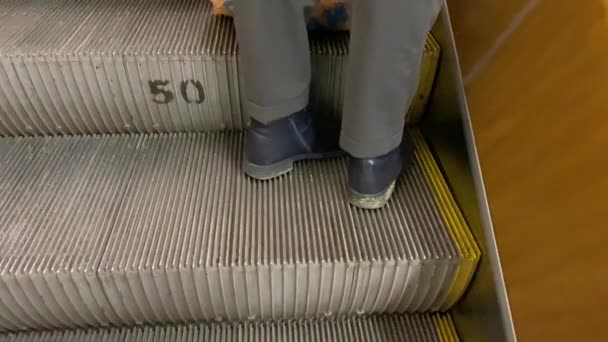 Los pies en pantalones y botas de zapatos azules sucios están en el escalón de la escalera mecánica. Movimiento automático de peatones a diferentes alturas. Transporte de pasajeros metro en la estación de tren. Concepto de viaje de negocios 4k — Vídeos de Stock