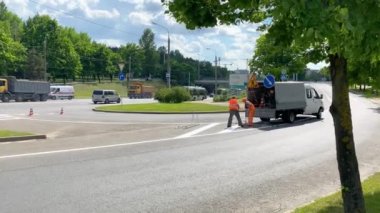 Parlak güvenlik üniformalı el işçisi yol işaretleme makinesini asfalt kıvrımında ısıtmalı boya ile beyaz çizgi boyuyor. Bir araba geçiyor. Şehir caddesinde asfaltta. 02.16.2021 Minsk Belarus 4k