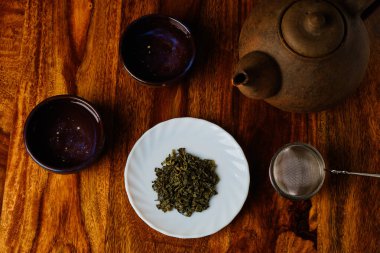 Beyaz tabakta yeşil çay ve süzgeç, tahta masa manzaralı kâseli çaydanlık.