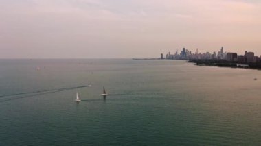 Eğlence amaçlı motorlu tekne, Michigan Gölü 'ne doğru yol alan birkaç yelkenlinin yanından geçerken Chicago ufuk çizgisi ve ufukta yükselen binaların önünden geçerken hava görüntüsü...