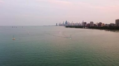 Gün batımından önce Chicago ufuk çizgisi ve ufukta yükselen binalarla birlikte yelkenlilerin yüzdüğü sakin Michigan Gölü 'nde kayarak geçen bir motorlu tekne görüntüsü..