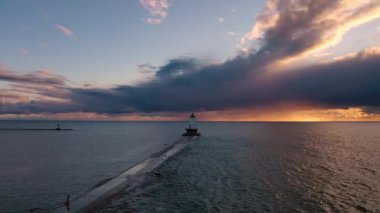 Dalgalar yükselirken dalgaların ötesindeki mavi ve turuncu renkli bulutların önünde beyaz renkli deniz feneriyle dalgaların üzerinde güzel bir günbatımı havası süzülüyor..