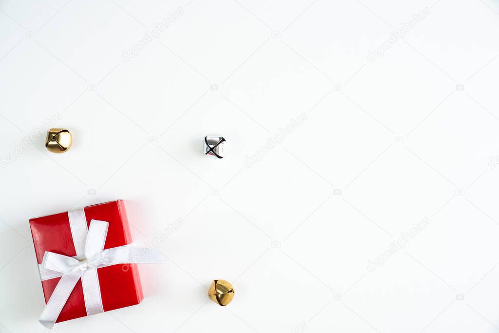 Beautiful holiday or Christmas background image of a small white ribbon wrapped red box present or gift in the lower left corner on a white backdrop with silver and gold jingle bells and copy space.