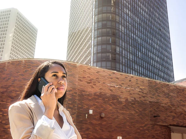 Mixed race African American business woman wearing a beige colored blazer and white shirt stands outside of a glass and steel highrise building in the city as she takes a phone call on her cell phone.