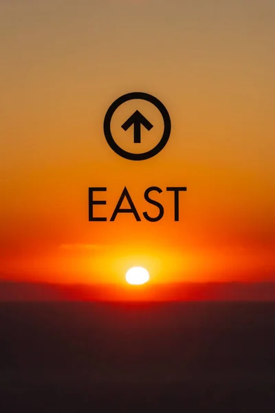 A bright yellow sun rises over the water of Lake Michigan directly to the east in the distance below a graphic and arrow on a window pointing out the direction of the view with orange sky.