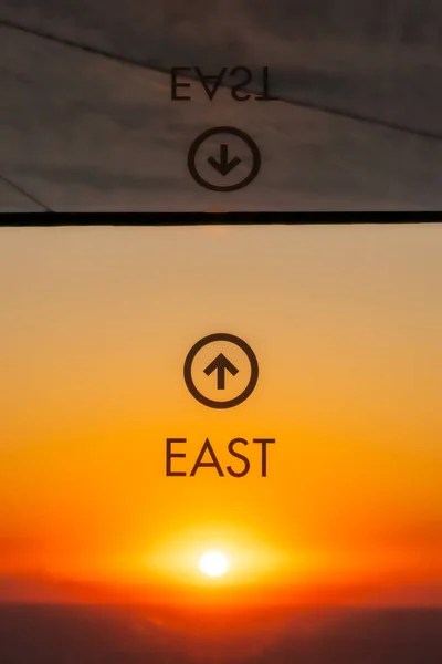 A bright yellow sun rises over the water of Lake Michigan directly to the east in the distance below a graphic and arrow on a window pointing out the direction of the view with orange sky.