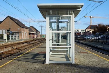 Boş bir tren istasyonu platformunda modern cam bir sığınak ve paralel raylar bulunur. Parlak mavi gökyüzünün altında binalar ve arka planda endüstriyel bir vinç bulunur..