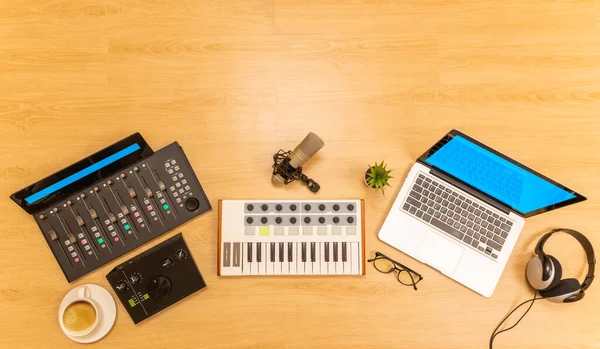 digital music production equipment. midi keyboard, audio interface, control surface, condenser microphone, headphone, laptop computer on wooden desk