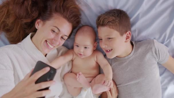 Çocuklarıyla mutlu bir anne, babasıyla veya akrabalarıyla bir yatakta selfie ya da video görüşmesi yapıyor. Teknoloji kavramı, yeni nesil, aile, bağlantı, ebeveynlik, gerçeklik. — Stok video