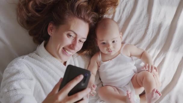 Close-up van moeder en haar pasgeboren baby het maken van een selfie of video-oproep naar vader of familie in een bed. Concept van technologie, nieuwe generatie, familie, verbinding, ouderschap. — Stockvideo