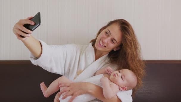 Anne ve yeni doğmuş bebeğinin bir yatakta babaya ya da akrabalara selfie ya da video görüşmesi yaparken yakın çekim. Teknoloji kavramı, yeni nesil, aile, bağlantı, ebeveynlik. — Stok video