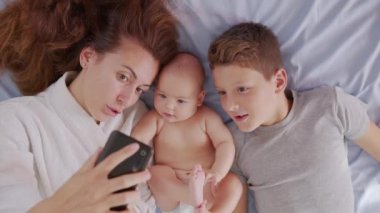 Çocuklarıyla mutlu bir anne, babasıyla veya akrabalarıyla bir yatakta selfie ya da video görüşmesi yapıyor. Teknoloji kavramı, yeni nesil, aile, bağlantı, ebeveynlik, gerçeklik.