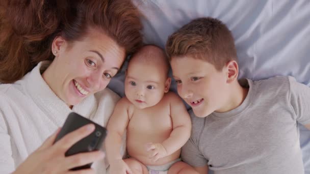 子供たちと幸せな母親は、ベッドの中で父親や親戚にセルフィーやビデオ通話をしています。テクノロジー、新世代、家族、つながり、親、真正性の概念. — ストック動画