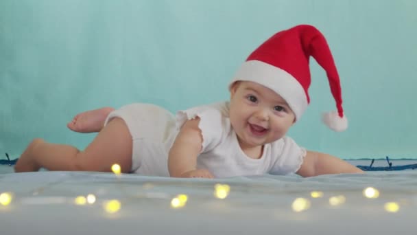 Lustigt litet barn som ler. Söta spädbarn pojke bär Santa hatt liggande på soffan. — Stockvideo