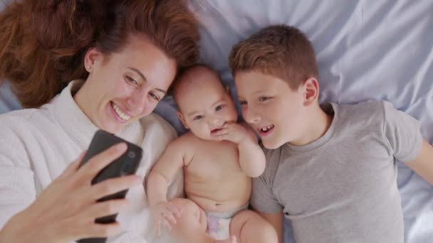 La madre feliz con sus hijos está haciendo una autofoto o una videollamada a su padre o familiares en una cama. Concepto de tecnología, nueva generación, familia, conexión, paternidad, autenticidad. — Vídeo de stock