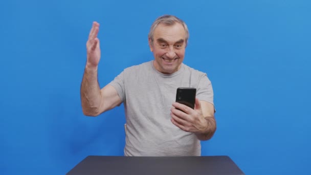 Happy grandfather winner mobile app sport bet holding smartphone having positive emotion. Overjoyed elderly man laughing celebrating victory success read good news message. Isolated on blue. — Stock Video