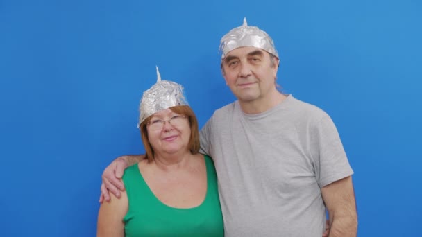 Ένα ηλικιωμένο ζευγάρι με προστατευτικά καπέλα από 5g ακτινοβολίας που στέκεται σε μπλε φόντο αγκαλιάζοντας. — Αρχείο Βίντεο