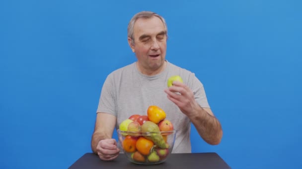 애플을 먹는 노인 남성 연금 수령자의 초상화. 머리에 회색 반점이 있고 카메라와 부드럽게 미소를 짓는 아름다운 남자. 건강 한 채식을 즐기 세요. 고립되어 있는 푸른. — 비디오