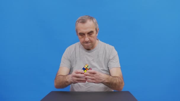 Літній чоловік, який вирішує Рубік. Ізольовані на синьому. — стокове відео