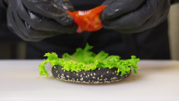 Haciendo hamburguesas. Las manos del chef en guantes negros ponen ensalada de lechuga verde fresca en el pan redondo de hamburguesa con salsa. Set en Negro brillante mesa reflectante. Vista de cerca. — Vídeos de Stock