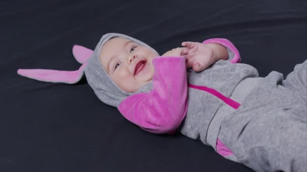 Μικρό παιδί με στολή πασχαλινού κουνελιού, κορυφαία θέα. Ένα ωραίο μικρό καυκάσιο νεογέννητο μωρό είναι αστείο χαμογελαστό, ξαπλωμένο πίσω στην κούνια του παιδιού. Χαριτωμένο μωρό χαμογελά και κοιτάζοντας στην κάμερα από κοντά. — Αρχείο Βίντεο