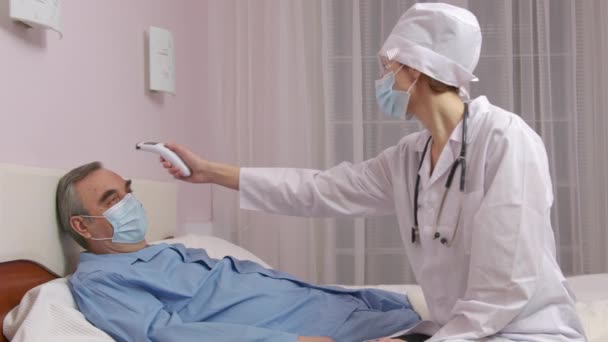 Gros plan d'une femme médecin caucasienne mesurant à la main la température d'un homme d'âge moyen couché sur un lit souffrant de Covid-19 à l'aide d'un thermomètre numérique infrarouge. — Video