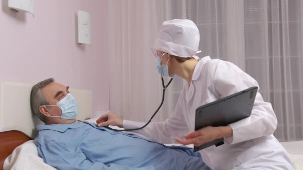 Il medico femminile usa lo stetoscopio per ascoltare il cuore del paziente anziano. Visita a domicilio dell'infermiera, controllo dello stato di salute del vecchio durante il coronavirus, quarantena covid-19. — Video Stock