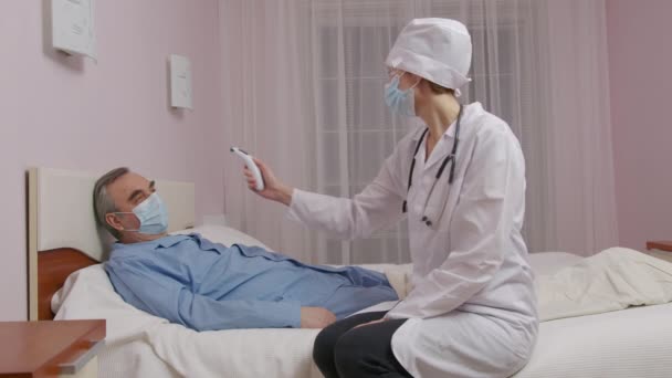 Primer plano de la mano del médico caucásico que mide la temperatura del paciente masculino de mediana edad acostado en la cama que sufre de Covid-19 usando termómetro digital infrarrojo. — Vídeos de Stock