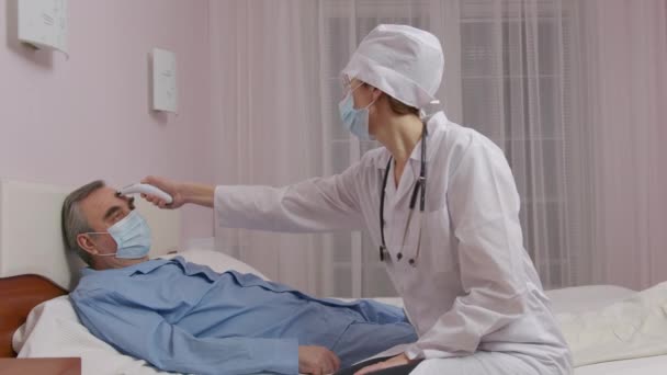 Zbliżenie białej kobiety lekarz ręka pomiar temperatury w średnim wieku męski pacjent leżący na łóżku cierpiący na Covid-19 za pomocą termometru cyfrowego podczerwieni. — Wideo stockowe