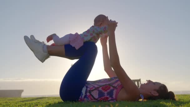 Šťastná rodina mladá maminka zvedání roztomilé malé dítě dcera nahoru létat jako letadlo baví venku na trávě v blízkosti oceánu. — Stock video