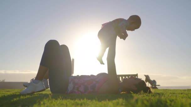 Jovem mãe está mantendo em seus braços e brincando com uma menina em uma praia oceânica ao pôr do sol durante as férias. — Vídeo de Stock