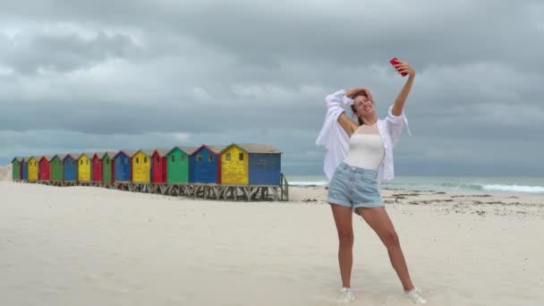 Een meisje maken selfie op het witte zand van Zuid-Afrika in de buurt van de kleurrijke bos Muizenberg Beach Houses. — Stockvideo