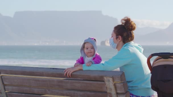 Kvinna med baby på stranden på bakgrundsbordet mountaine bär en skyddande ansiktsmask för att förhindra Covid-19 Coronavirus pandemi. — Stockvideo