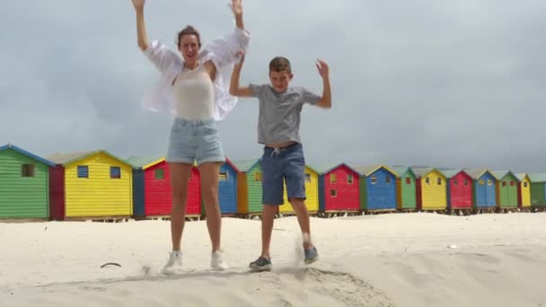 Moeder met zoon dansen op het witte zand van Zuid-Afrika in de buurt van het kleurrijke bos Muizenberg Beach Houses. — Stockvideo