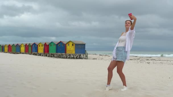 Una chica hace selfie en las arenas blancas de Sudáfrica cerca de la colorida madera Muizenberg Beach Houses. — Vídeos de Stock