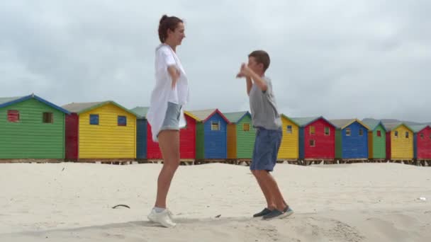 Een meisje met jongen dansen op het witte zand van Zuid-Afrika in de buurt van het kleurrijke bos Muizenberg Beach Houses. — Stockvideo